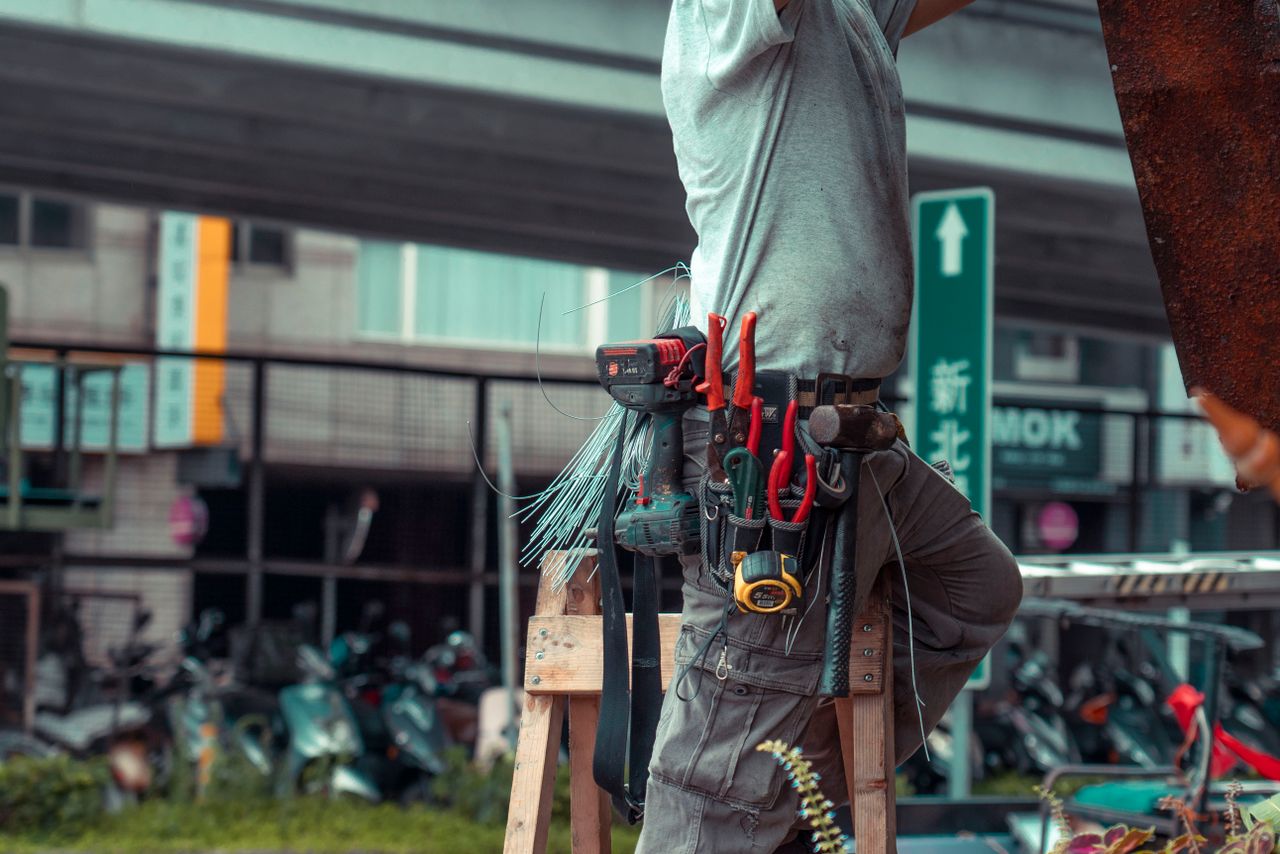 Handyman standing on a ladder with tool belt, repairing outdoors with visible tools.