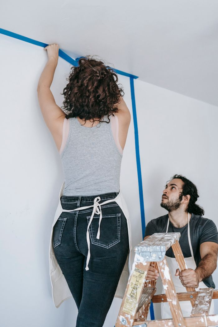 A couple working on a home renovation project using painter's tape and a ladder indoors.